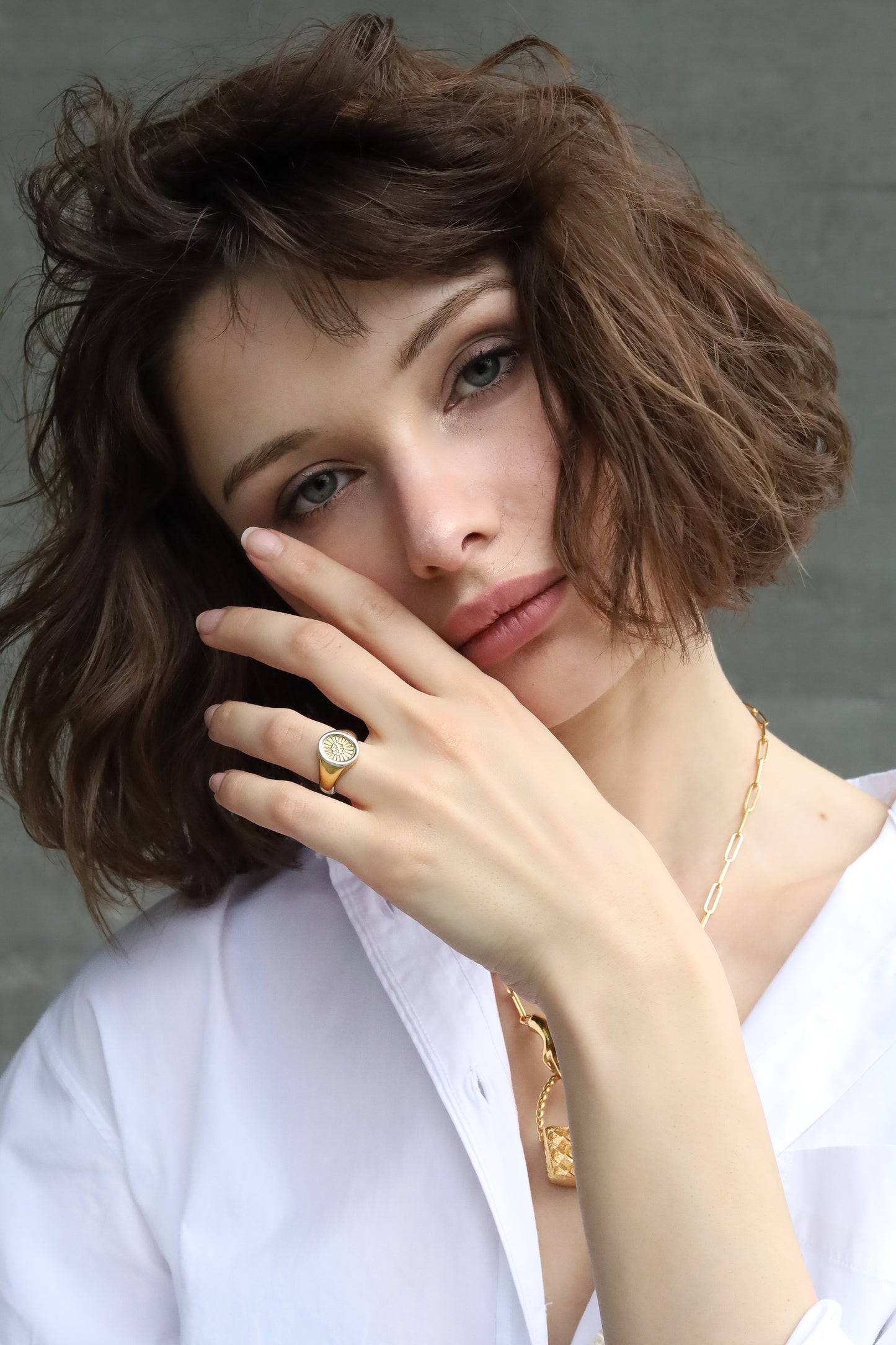 Woman with short brown hair wearing a gold ring and necklace against a neutral background