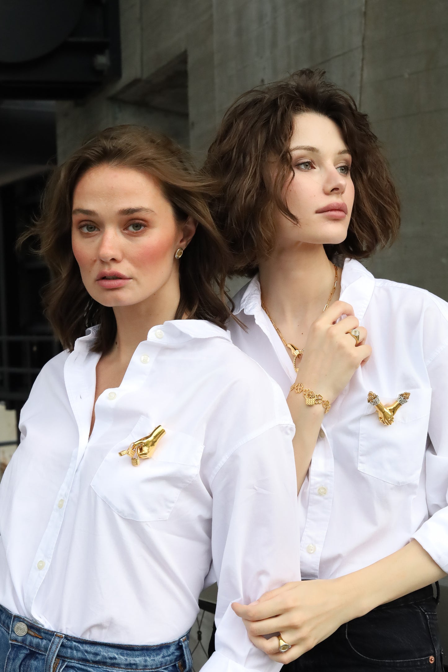 Two women wearing white shirts with gold Jenna London brooches in front of a gray wall.